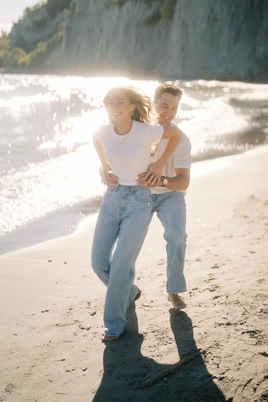 Young couple laughing and playing by the ocean on a sunny beach, wearing casual white shirts and jeans, with cliffs and sparkling water in the background.