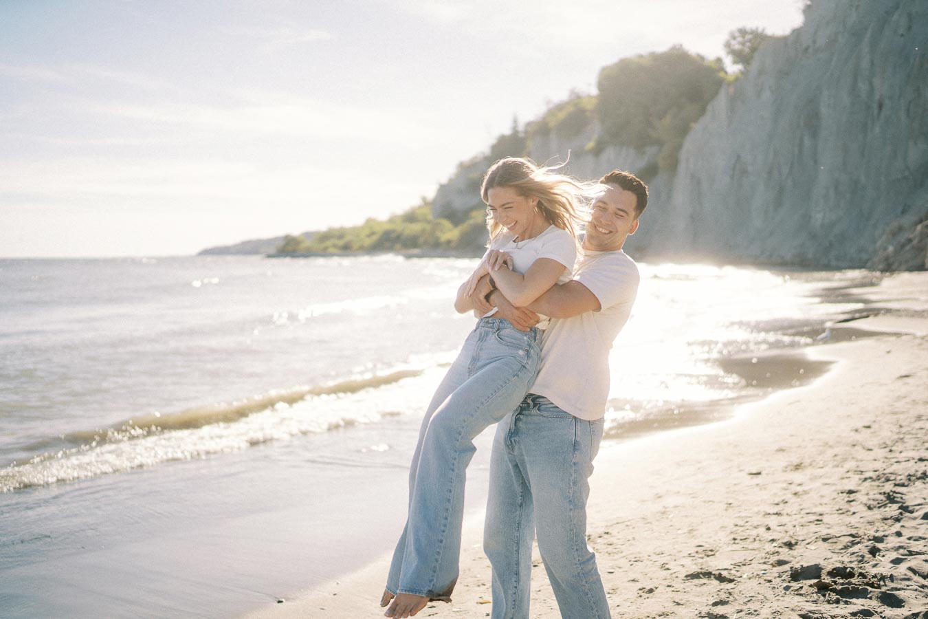 A happy couple enjoying a sunny day on a beach, with the man playfully lifting the woman while both laugh, wearing casual white shirts and jeans. The ocean waves gently lap the shore, with cliffs and greenery in the background.