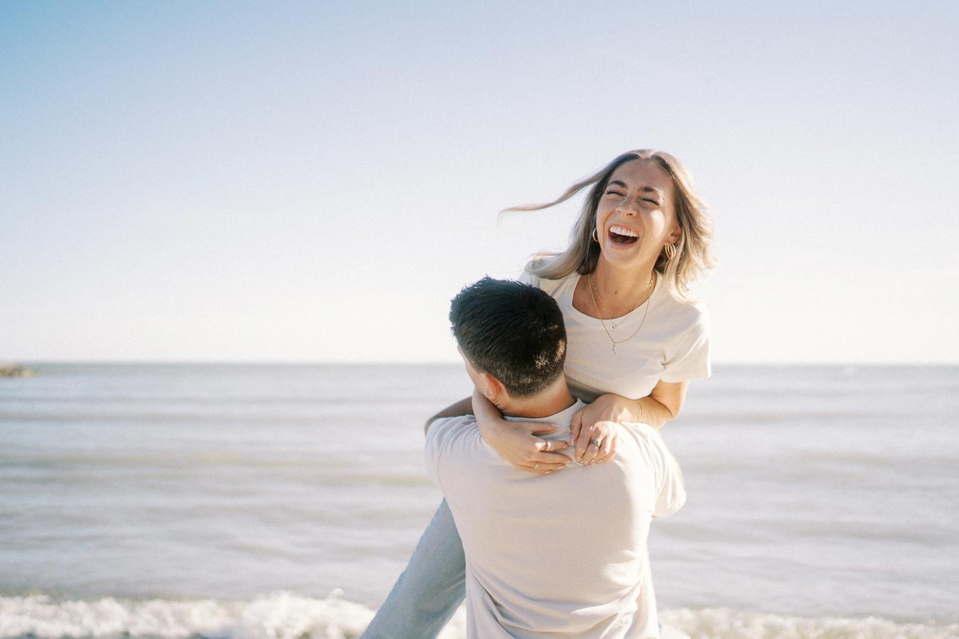 Young couple enjoying a playful moment by the beach, with a clear blue sky and ocean in the background. The woman is smiling happily while being lifted by her partner.