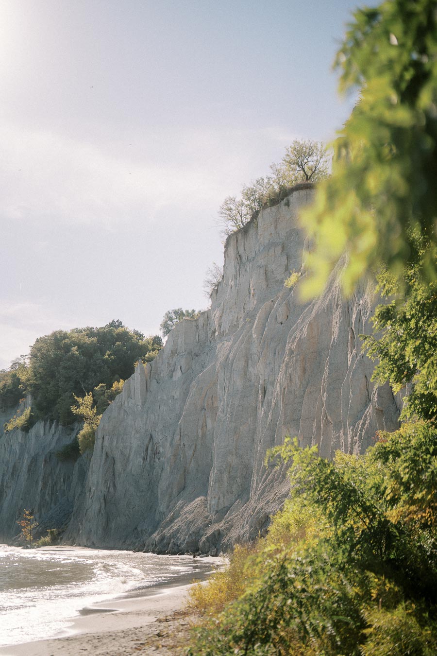 Scenic view of a coastal cliffside at Scarborough Bluffs, Toronto, surrounded by lush greenery and a sandy beach alongside the gentle waves of Lake Ontario.