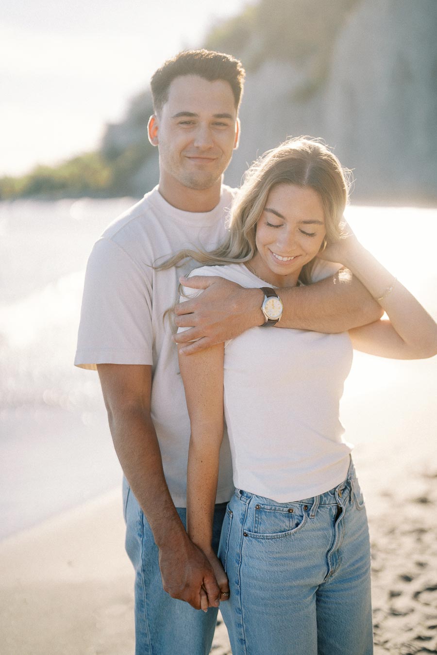 A couple in casual attire embracing and smiling on a sunlit beach, with the ocean and cliffs in the background, capturing a serene and romantic moment.