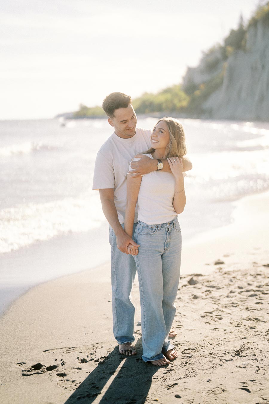 A couple embracing on a sunny beach with waves in the background, wearing casual white tops and jeans, enjoying a romantic moment by the seaside.