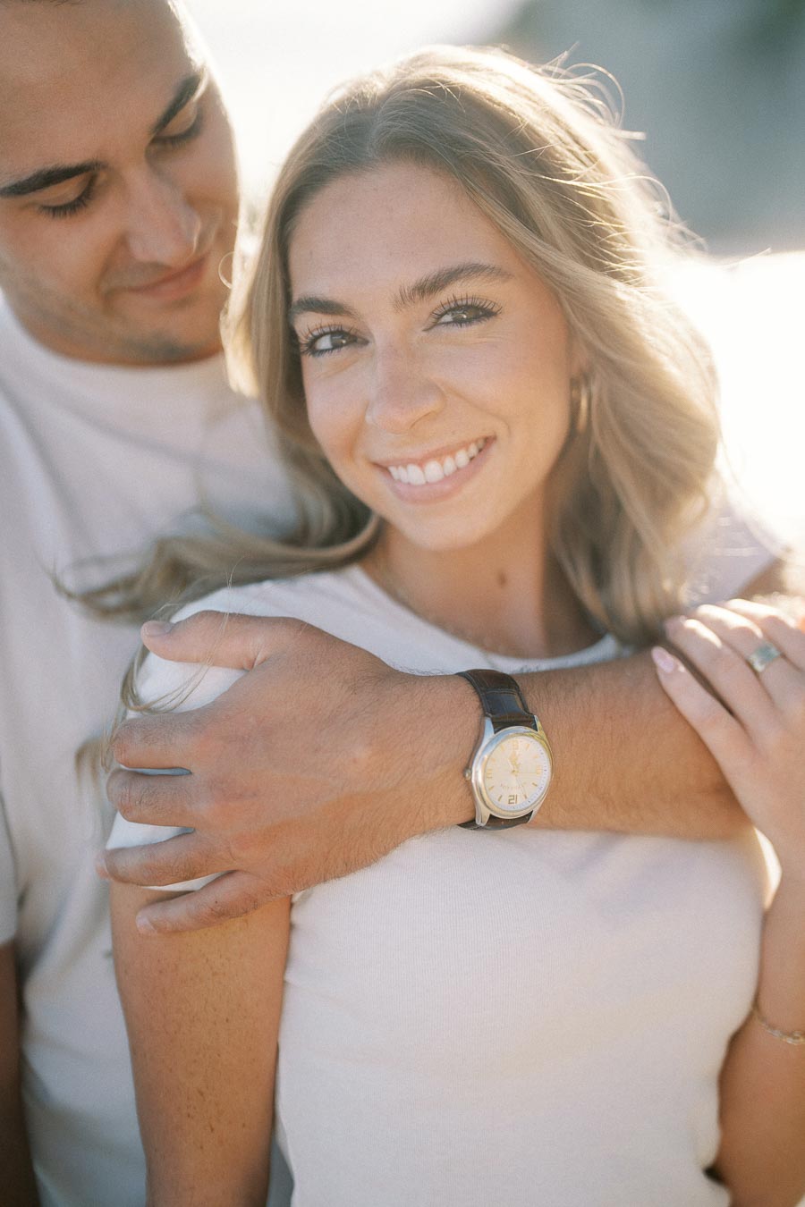 Smiling woman with long blonde hair embraced by a man, showcasing a vintage wristwatch, enjoying a sunny day outdoors.