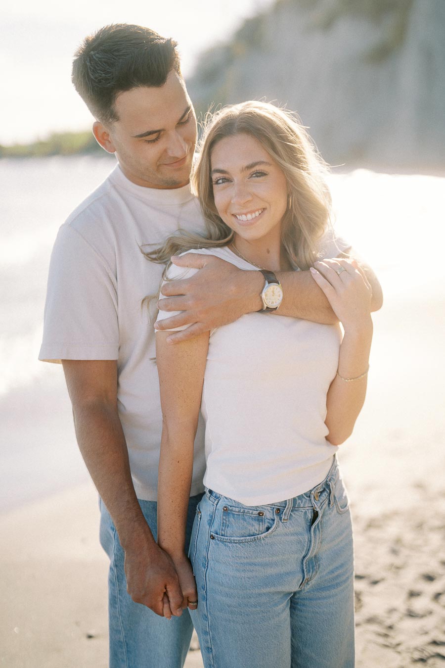 Happy couple embracing on a sunny beach, woman smiling at the camera while man holds her from behind, both wearing casual outfits.