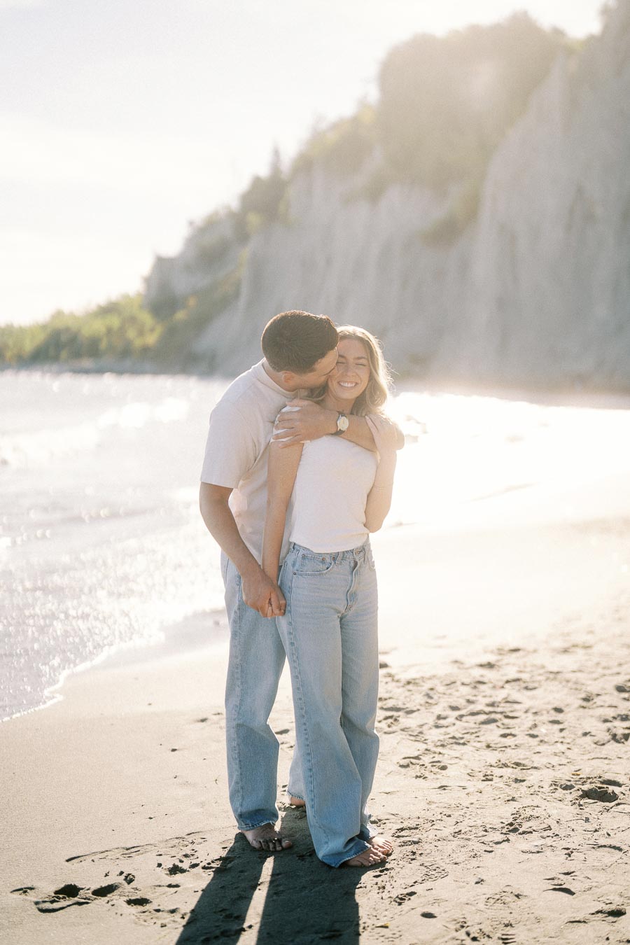 A happy couple embracing and smiling on a sunny beach, with cliffs and ocean waves in the background.