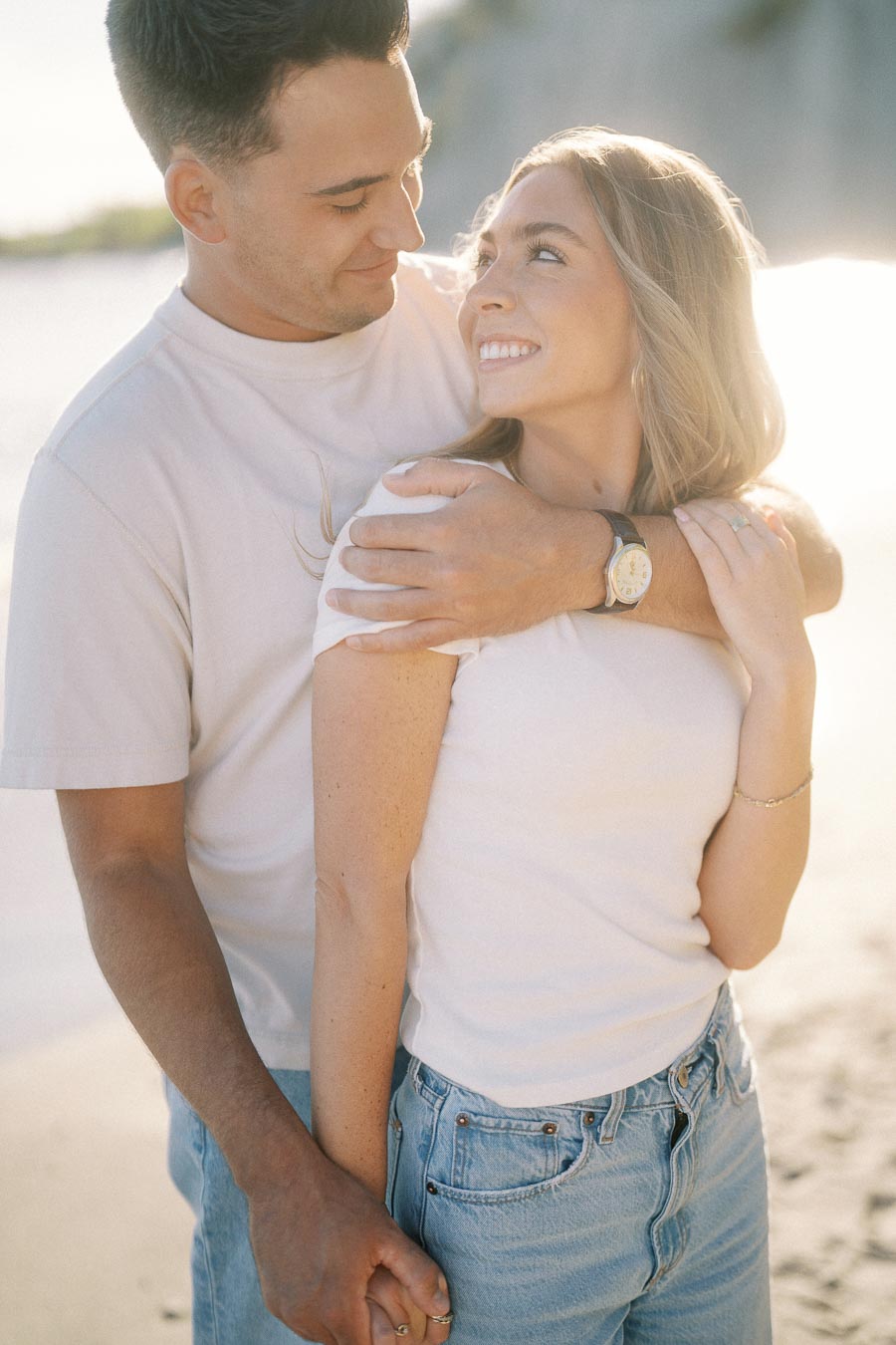 A couple smiling at each other while standing on a beach, embracing and holding hands, with soft sunlight in the background.