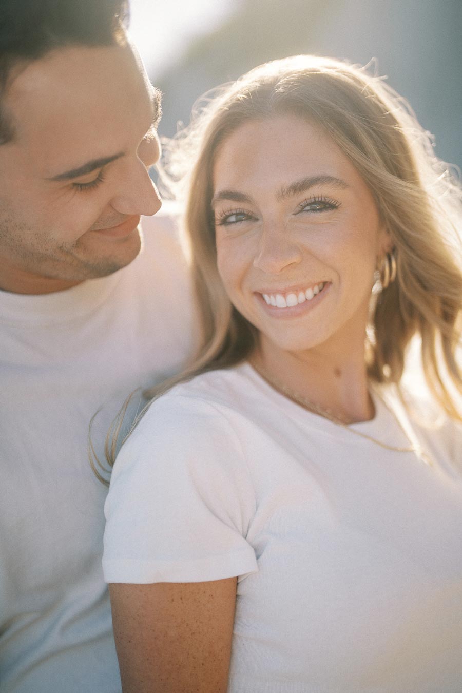 A happy couple enjoying a sunny day, with the woman smiling warmly and the man looking at her affectionately. Both wear casual white tops, conveying a sense of happiness and closeness.