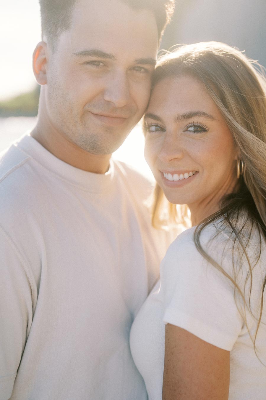 Couple smiling and embracing in natural sunlight, wearing casual white shirts, with a blurred outdoor background.