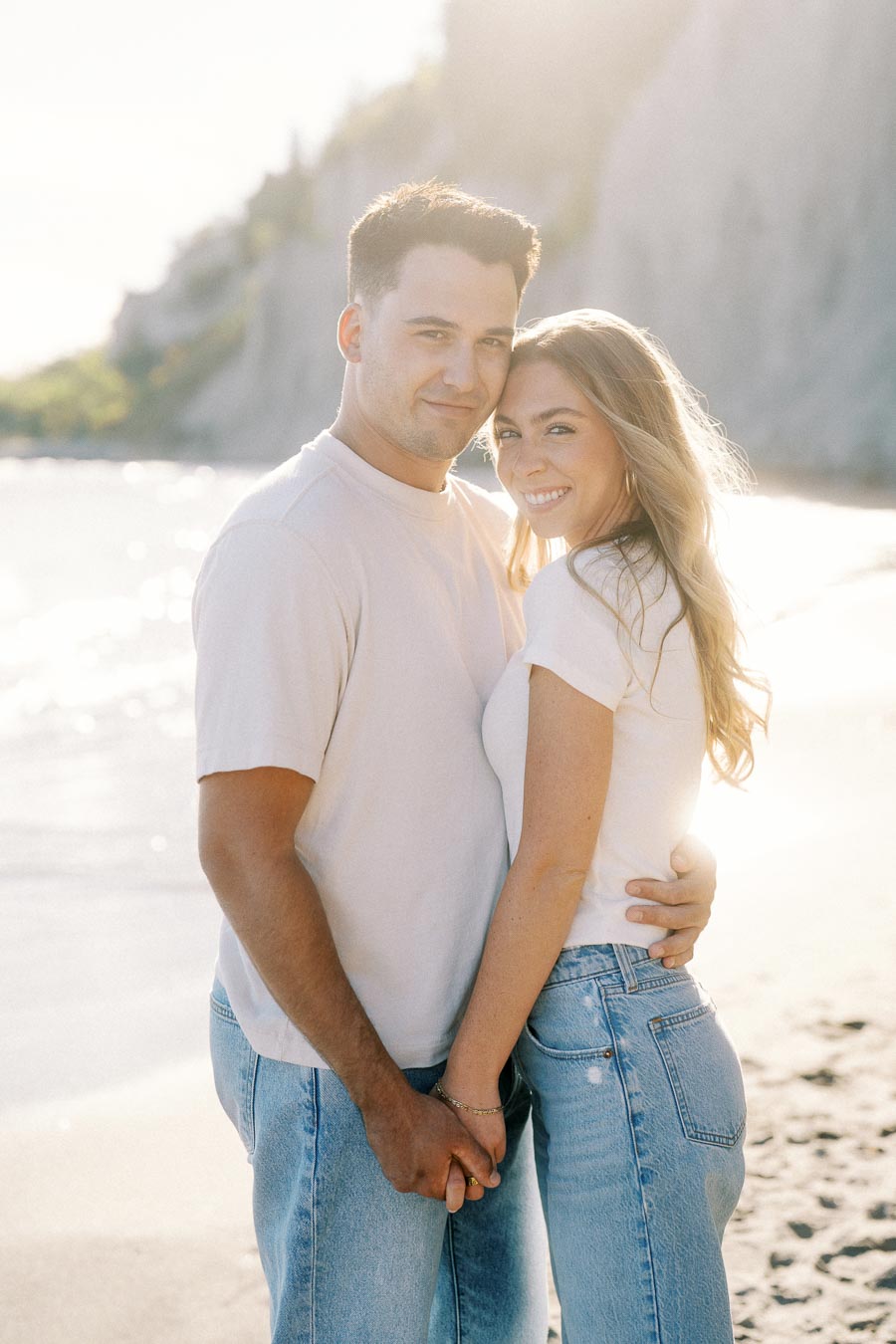 Smiling couple embracing on a sunlit beach, wearing casual outfits, with cliffs in the background.