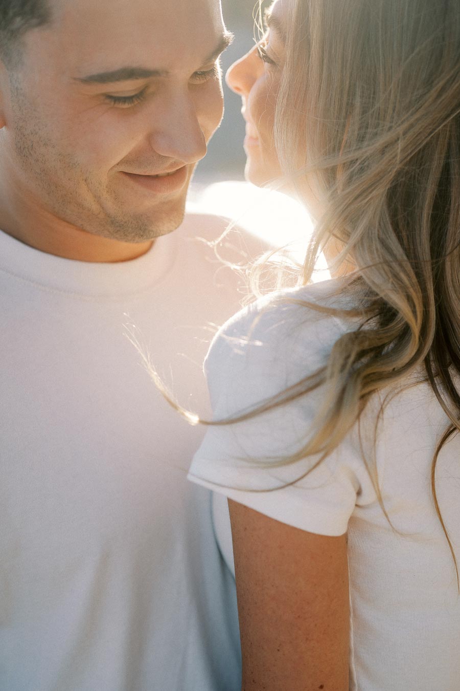 A couple gently smiling at each other, bathed in warm sunlight, wearing white shirts.