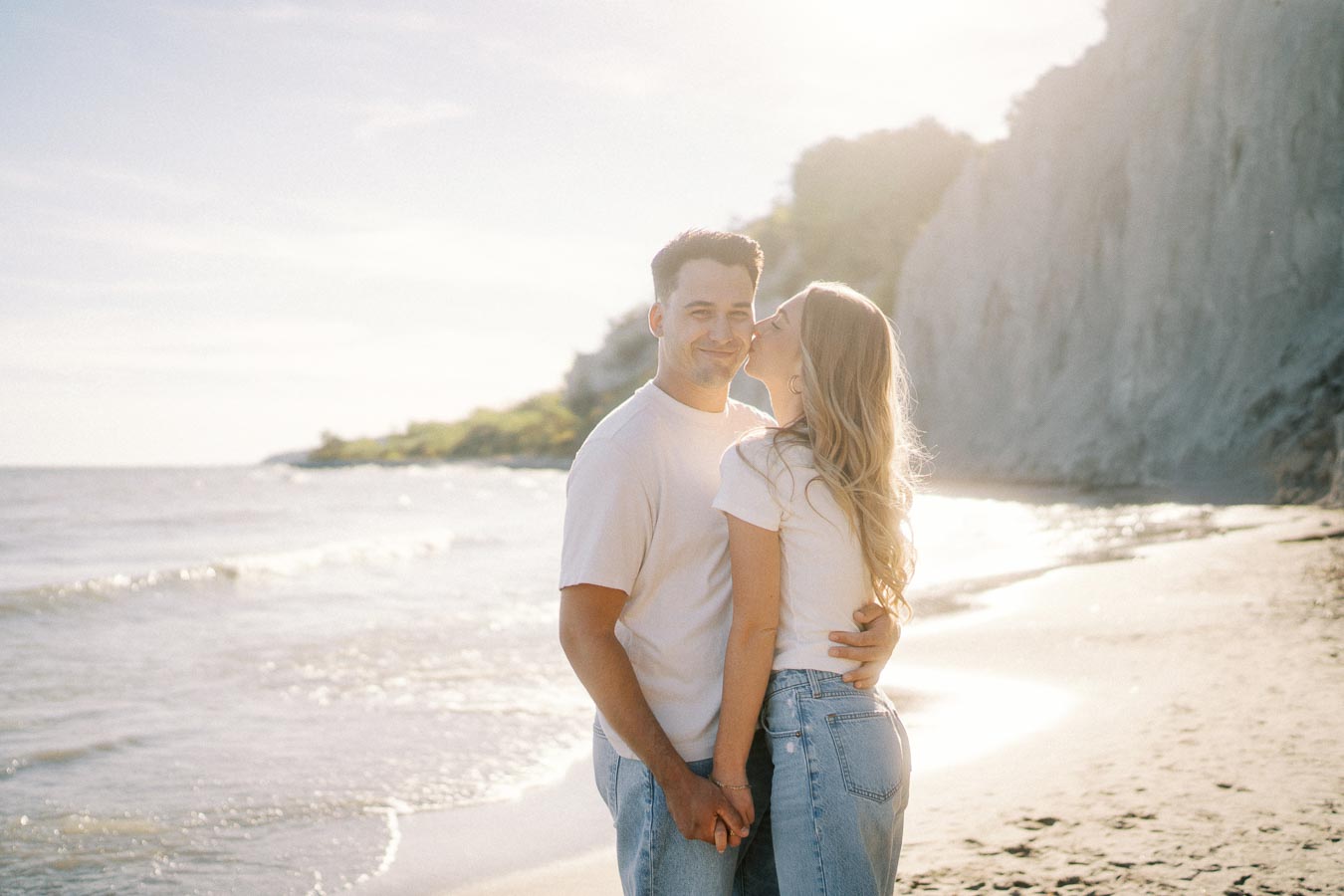 A couple embraces lovingly on a sunny beach, with the ocean and cliffs in the background, capturing a romantic and serene moment by the sea.
