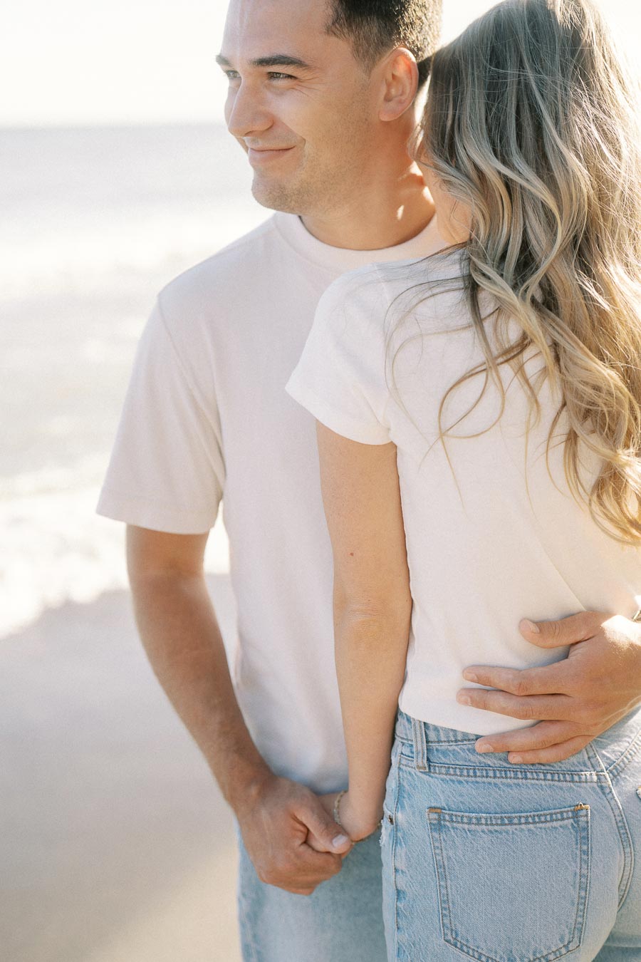 Happy couple holding hands and embracing on a sunny beach, dressed in casual white shirts and jeans, enjoying a romantic moment by the ocean.