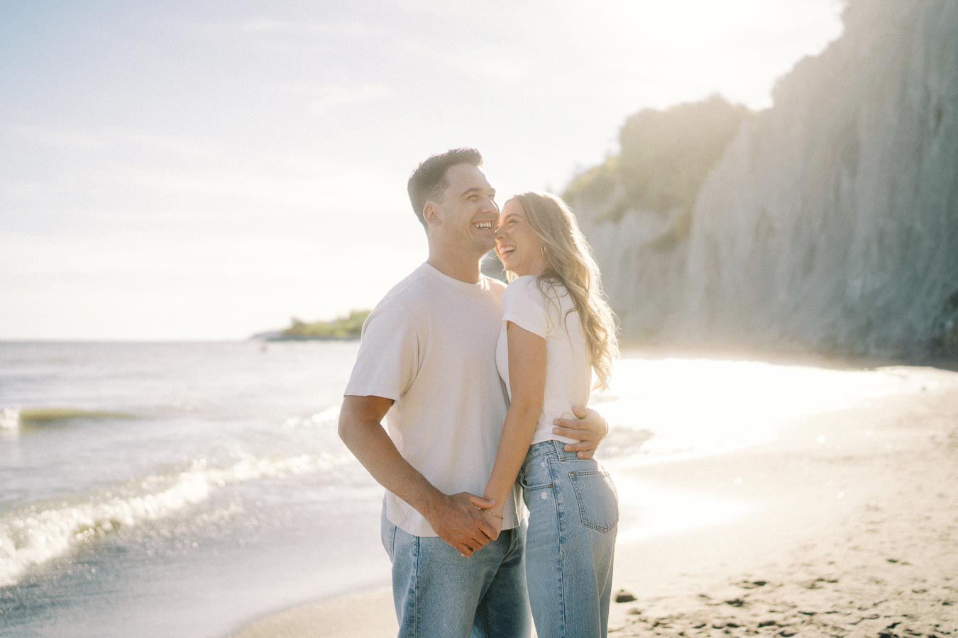 Couple smiling and embracing on a sunny beach, with waves gently crashing in the background and cliffs bathed in soft sunlight.