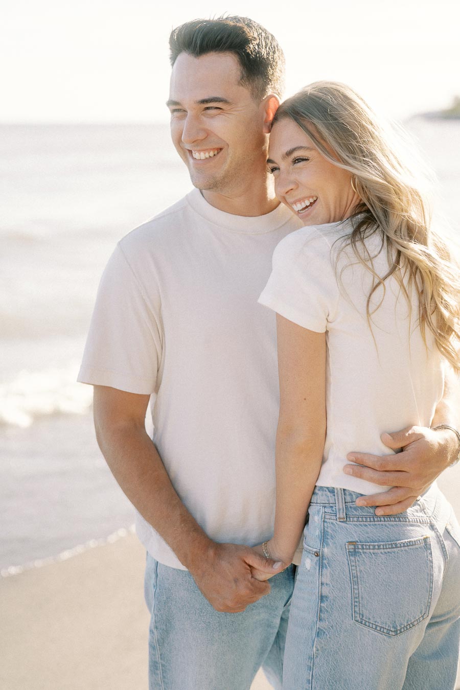 A joyful couple in casual attire, embracing and smiling on a sunny beach, enjoying their time together.