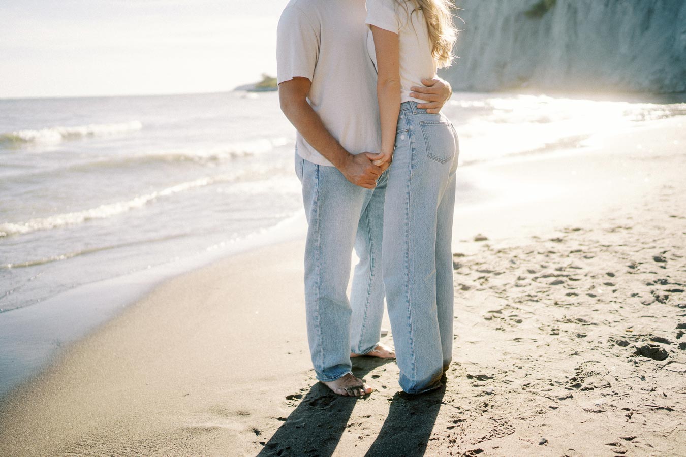 A couple in casual clothing, holding hands and facing the ocean on a sunny beach, with waves gently crashing in the background.