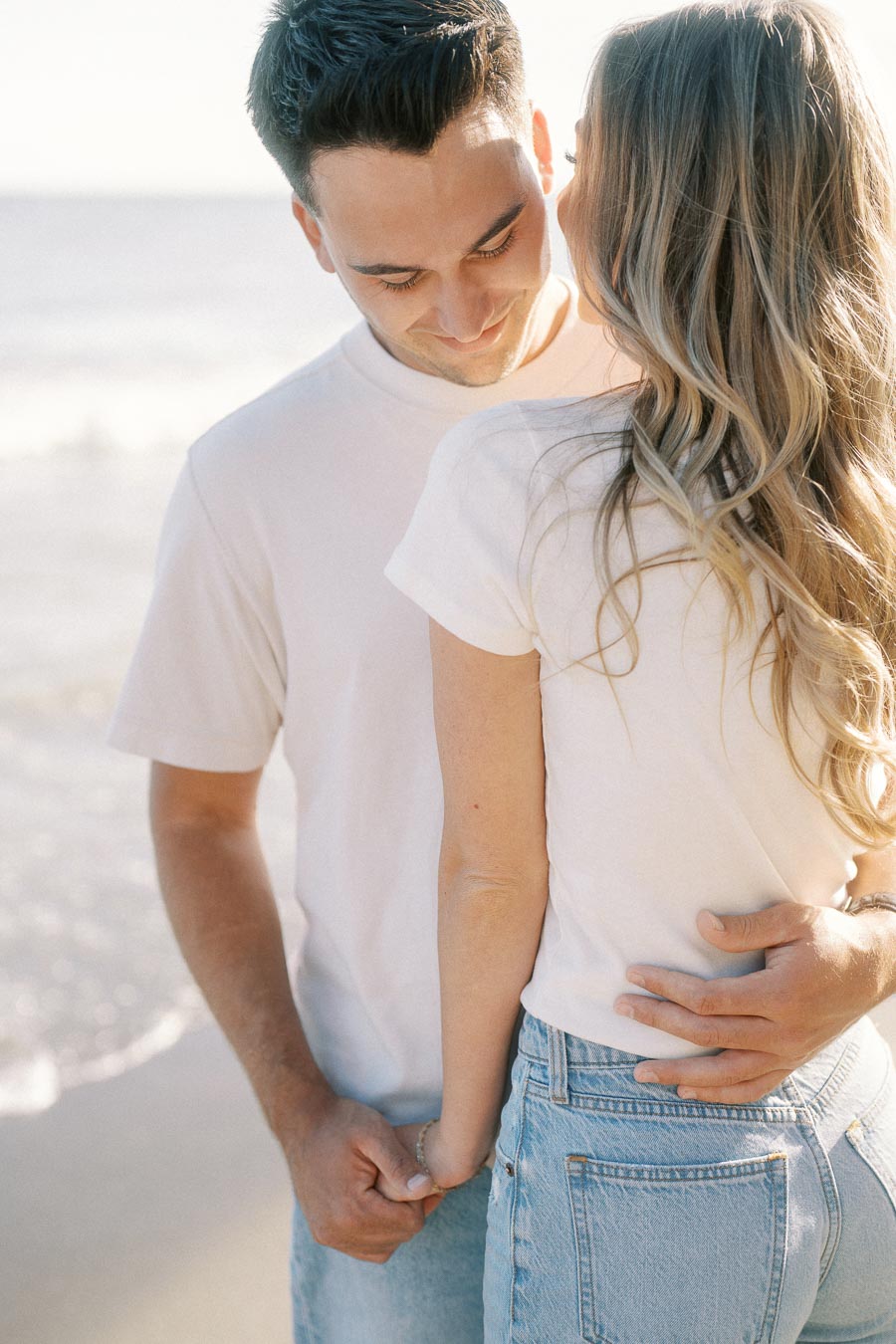 Young couple embracing on a sunny beach, wearing casual white shirts and jeans, with the ocean in the background.