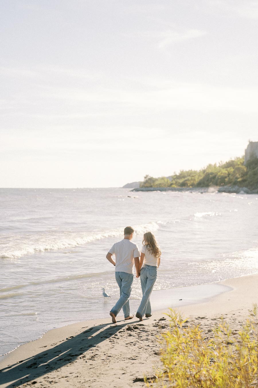 A couple holding hands walking along a sunny beach with gentle waves and distant cliffs, capturing a serene and romantic seaside moment.