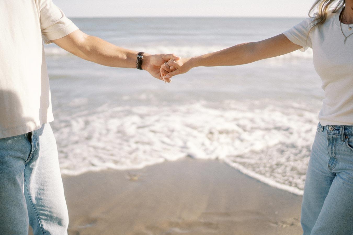 A couple holding hands on a sandy beach with gentle waves in the background, symbolizing connection and tranquility.