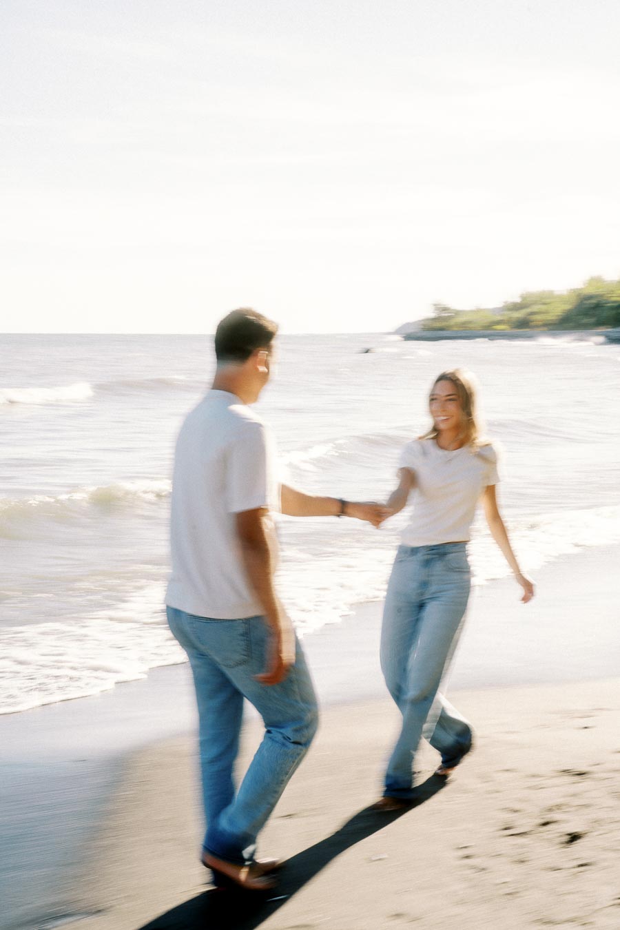 A blurred image of a couple holding hands and walking along a serene beach, with waves gently lapping at the shore on a sunny day.