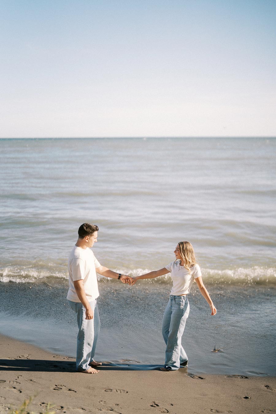 A couple holding hands and smiling on a sunny beach with waves in the background, both wearing casual jeans and white shirts, capturing a joyful moment by the ocean.