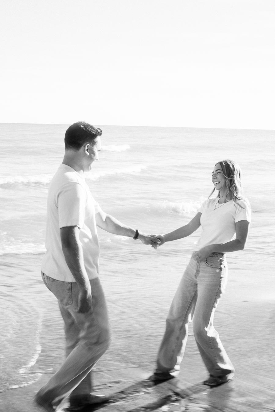 A cheerful couple holding hands and laughing while standing on a sandy beach, with waves gently rolling in the background.