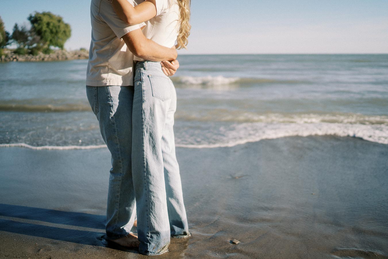 A couple embracing on a sandy beach with waves in the background, wearing casual jeans and light-colored shirts, under a clear blue sky.
