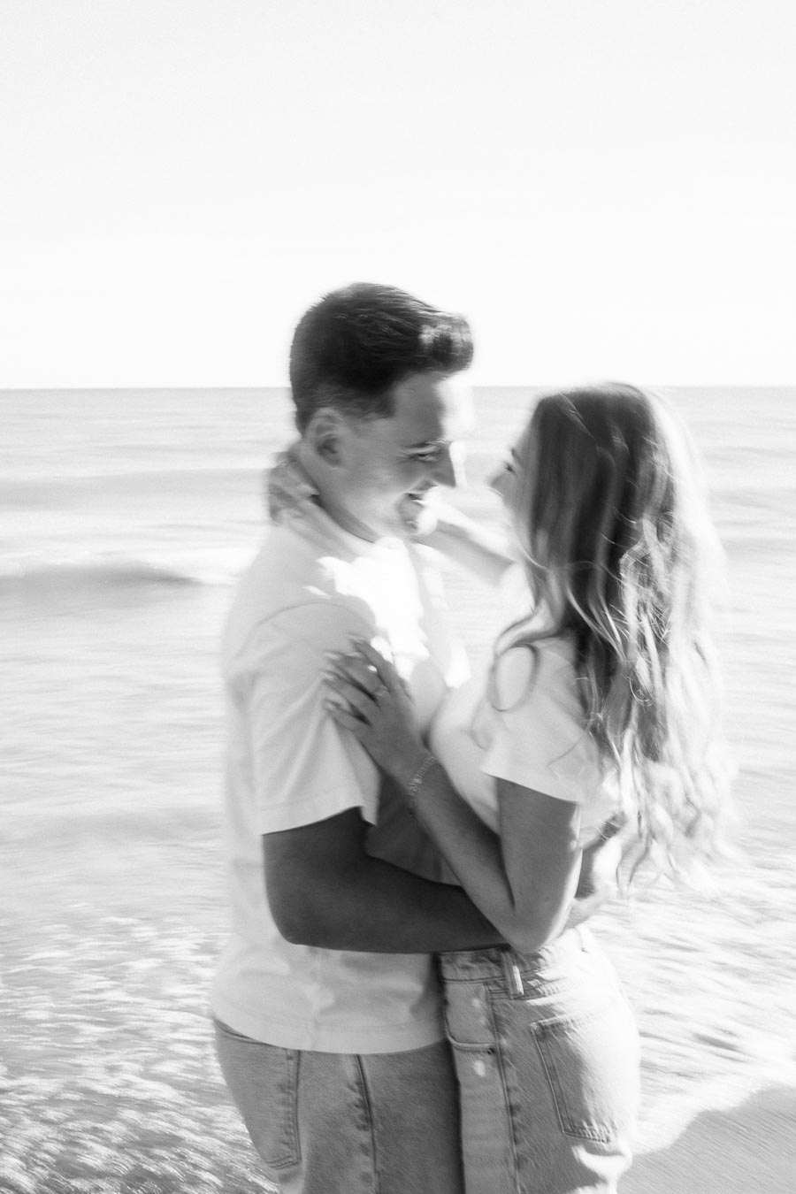 A black and white image of a couple embracing and smiling at each other on a beach, with the ocean in the background, creating a romantic and serene atmosphere.