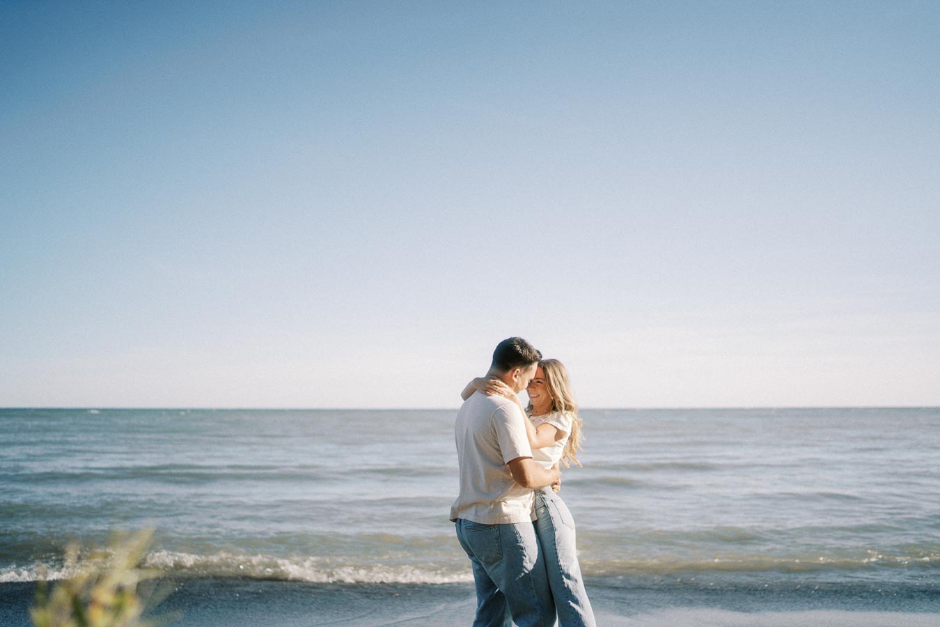 A couple embracing on a sunny beach, with clear blue skies and gentle ocean waves in the background.