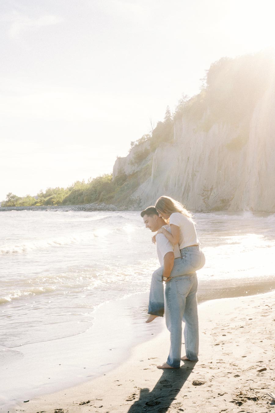 A couple enjoying a sunny day at the beach, with one giving the other a piggyback ride near the shoreline with cliffs in the background.