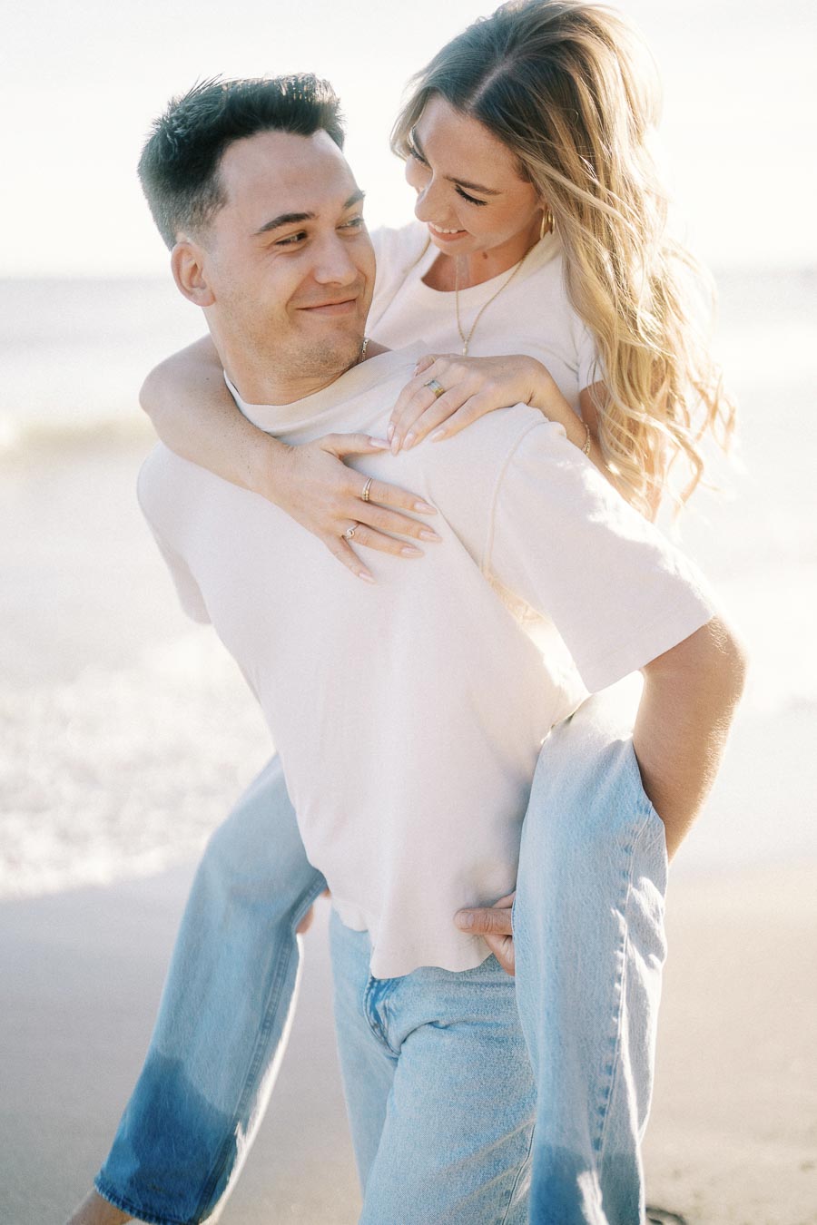 A couple laughing and enjoying a playful piggyback ride on a sunny beach, wearing casual jeans and white shirts, with the ocean and a clear sky in the background.