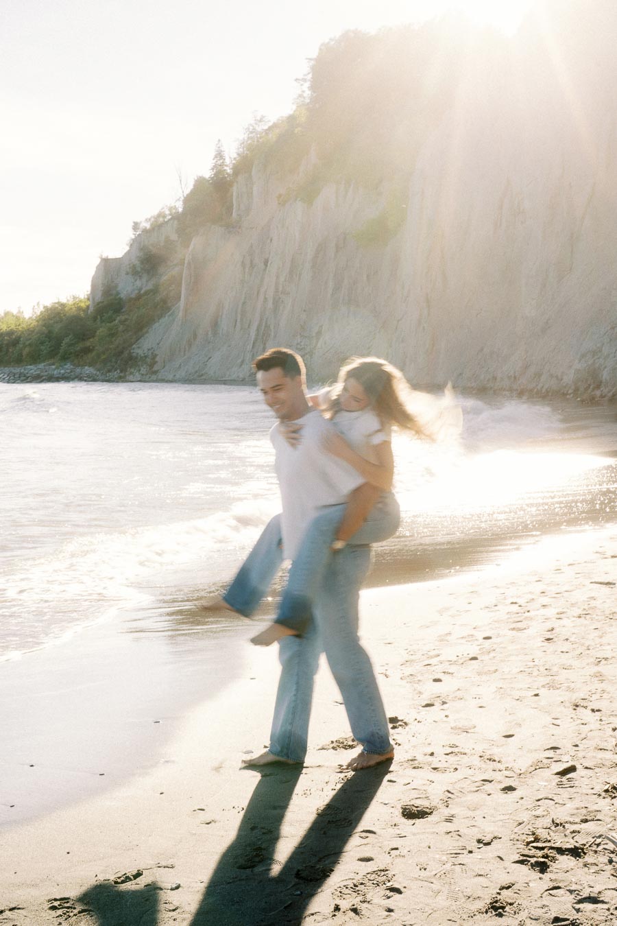 Couple enjoying a playful piggyback ride on a sunlit beach with cliffs in the background, capturing a romantic moment by the sea.