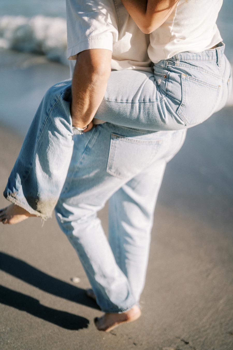A couple playfully walking on the beach, wearing light blue jeans and casual white shirts, with gentle ocean waves in the background.