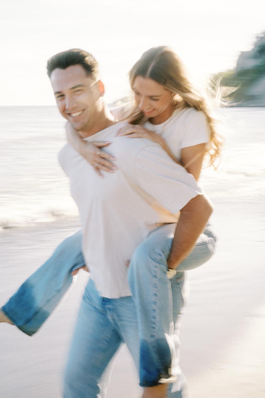 A couple enjoying a playful piggyback ride on a sandy beach at sunset, both wearing casual jeans and white shirts, with a serene ocean backdrop.