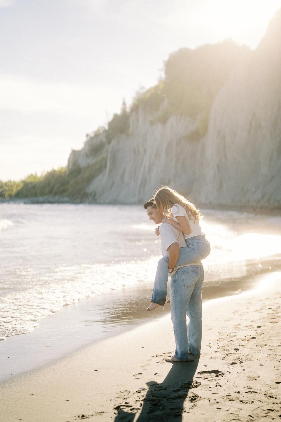 A joyful couple enjoying a playful piggyback ride on a sunny beach, with towering cliffs and a serene ocean in the background.