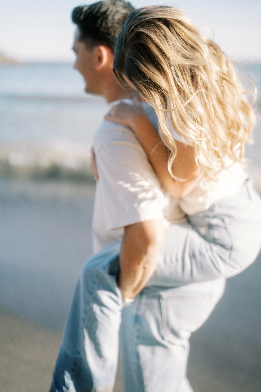 Romantic couple enjoying a playful piggyback ride on the beach, with ocean waves in the background and sunlight highlighting their relaxed poses and casual attire.