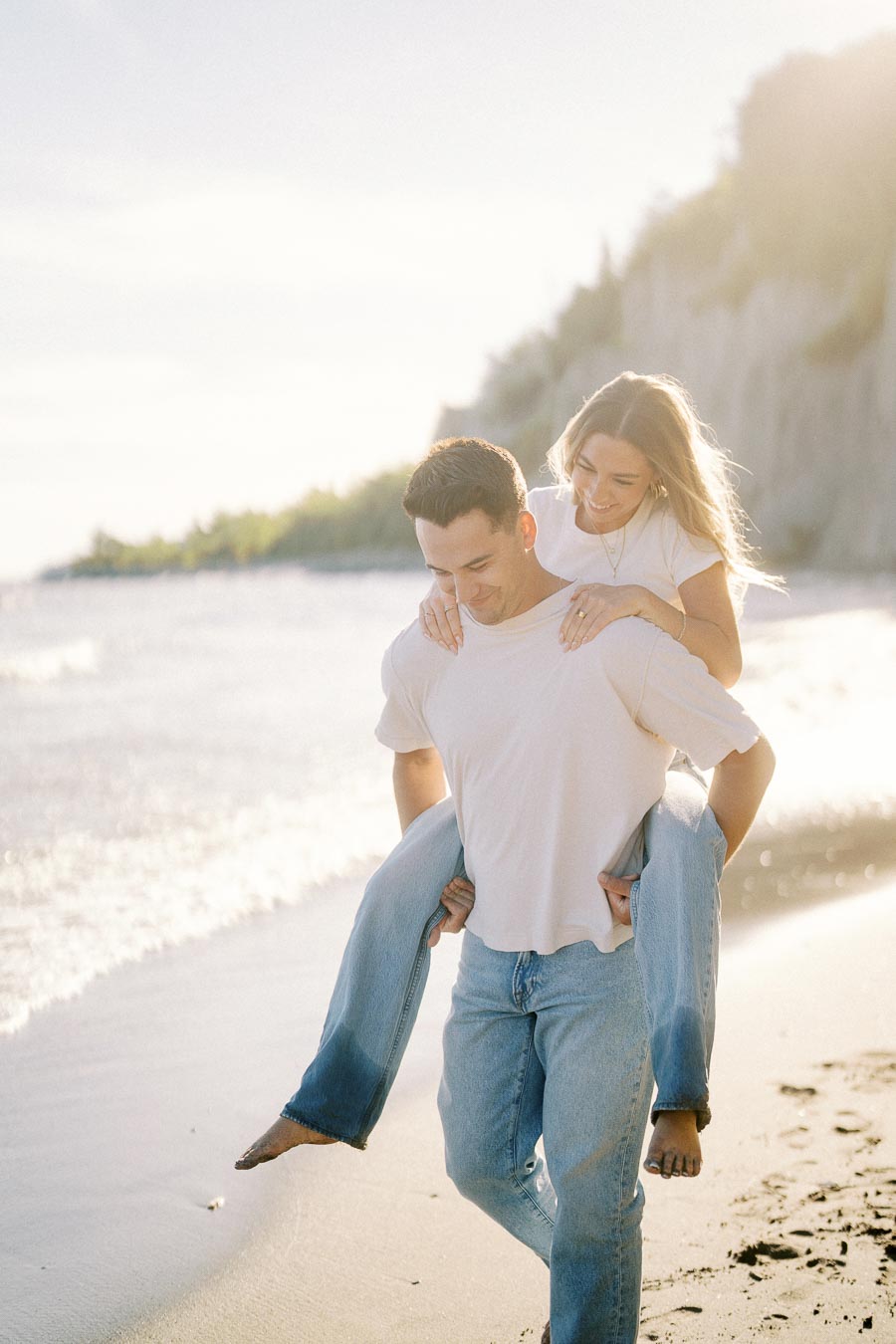 Young couple enjoying a playful piggyback ride on a sunny beach, both wearing casual white t-shirts and jeans, with scenic cliffs and ocean waves in the background.