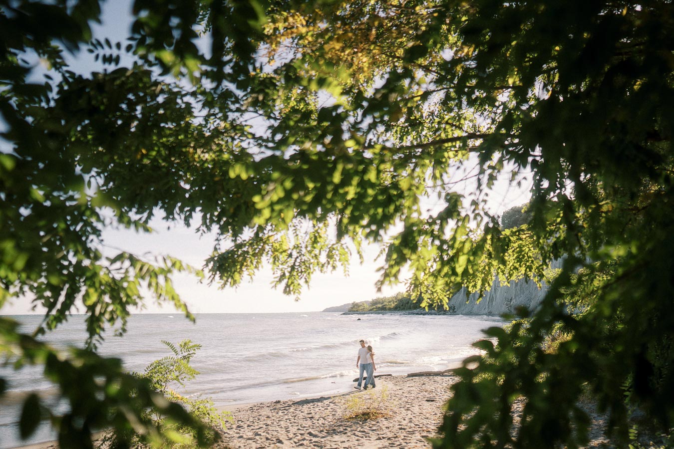 A couple walking along a scenic beach framed by lush green foliage on a sunny day, capturing a serene and romantic atmosphere by the water.