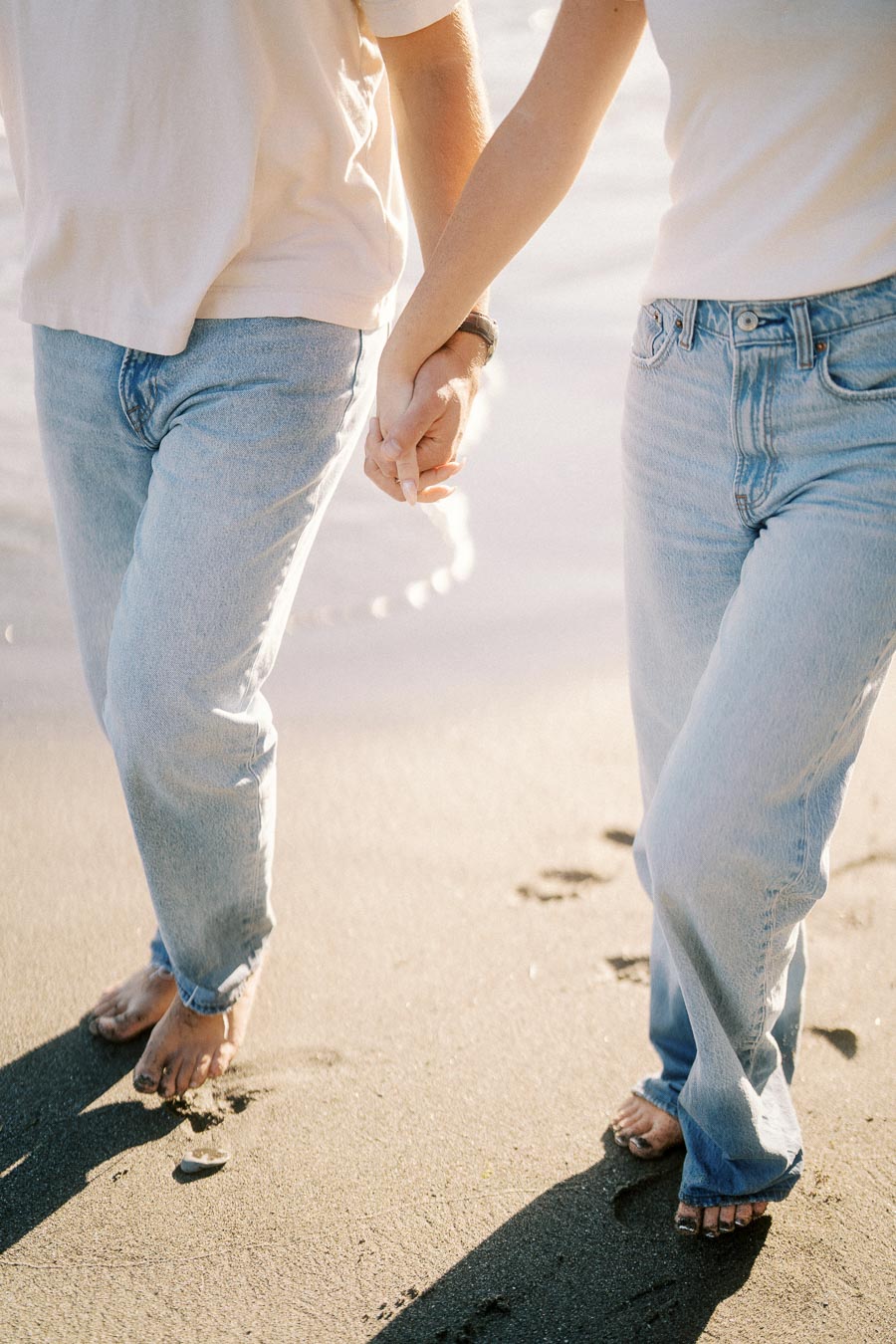 A couple holding hands while walking barefoot on a sandy beach, wearing casual light blue jeans and white shirts, with gentle waves in the background.
