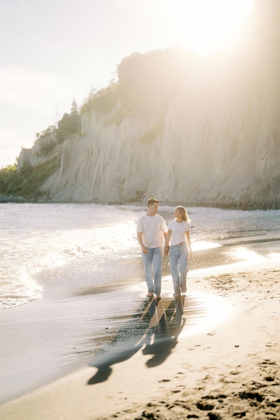 A couple walking hand in hand along a sunlit beach with scenic cliffs in the background, creating romantic shadows on the sand.