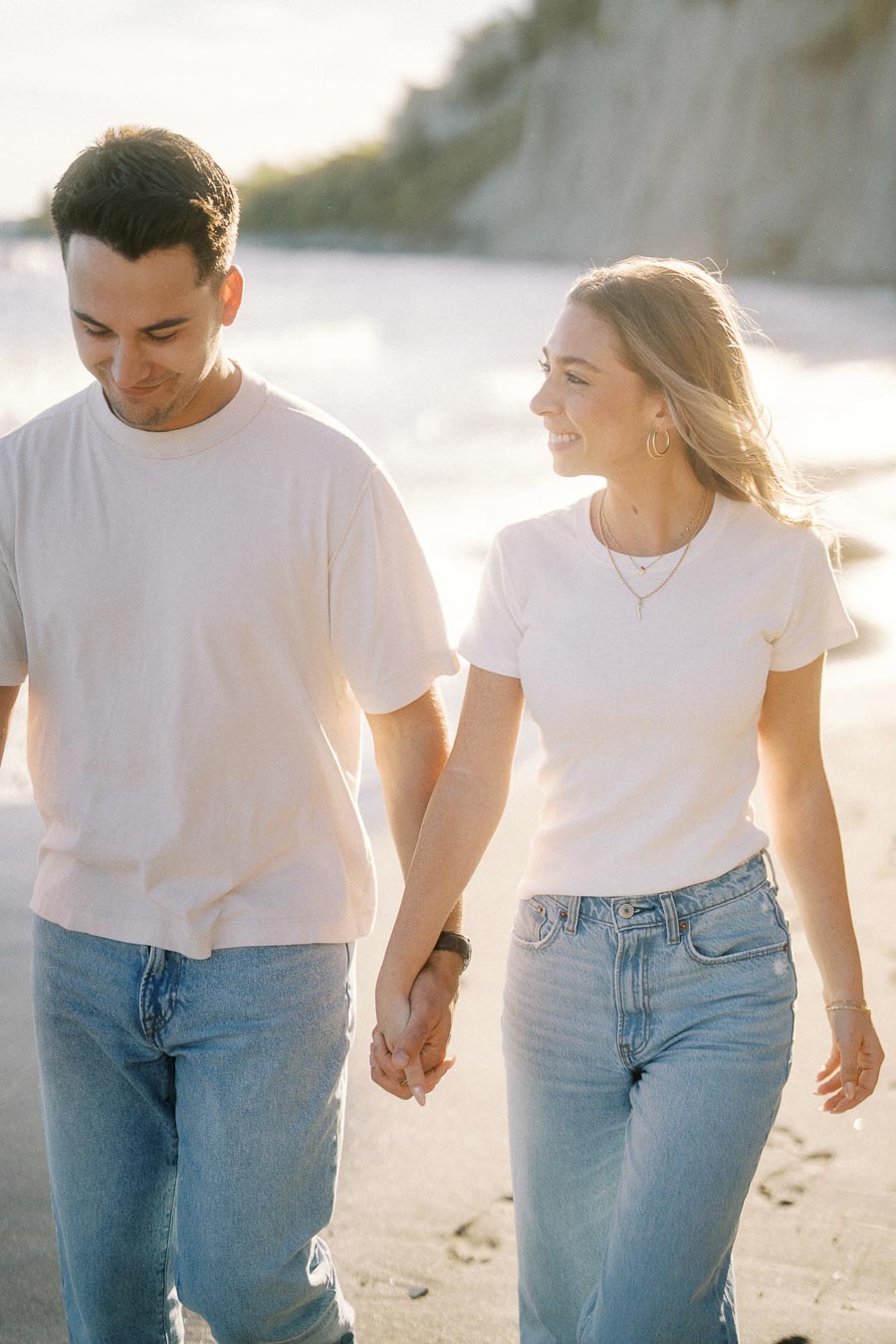 A couple holding hands and walking along a sunny beach, wearing casual matching outfits and smiling at each other.