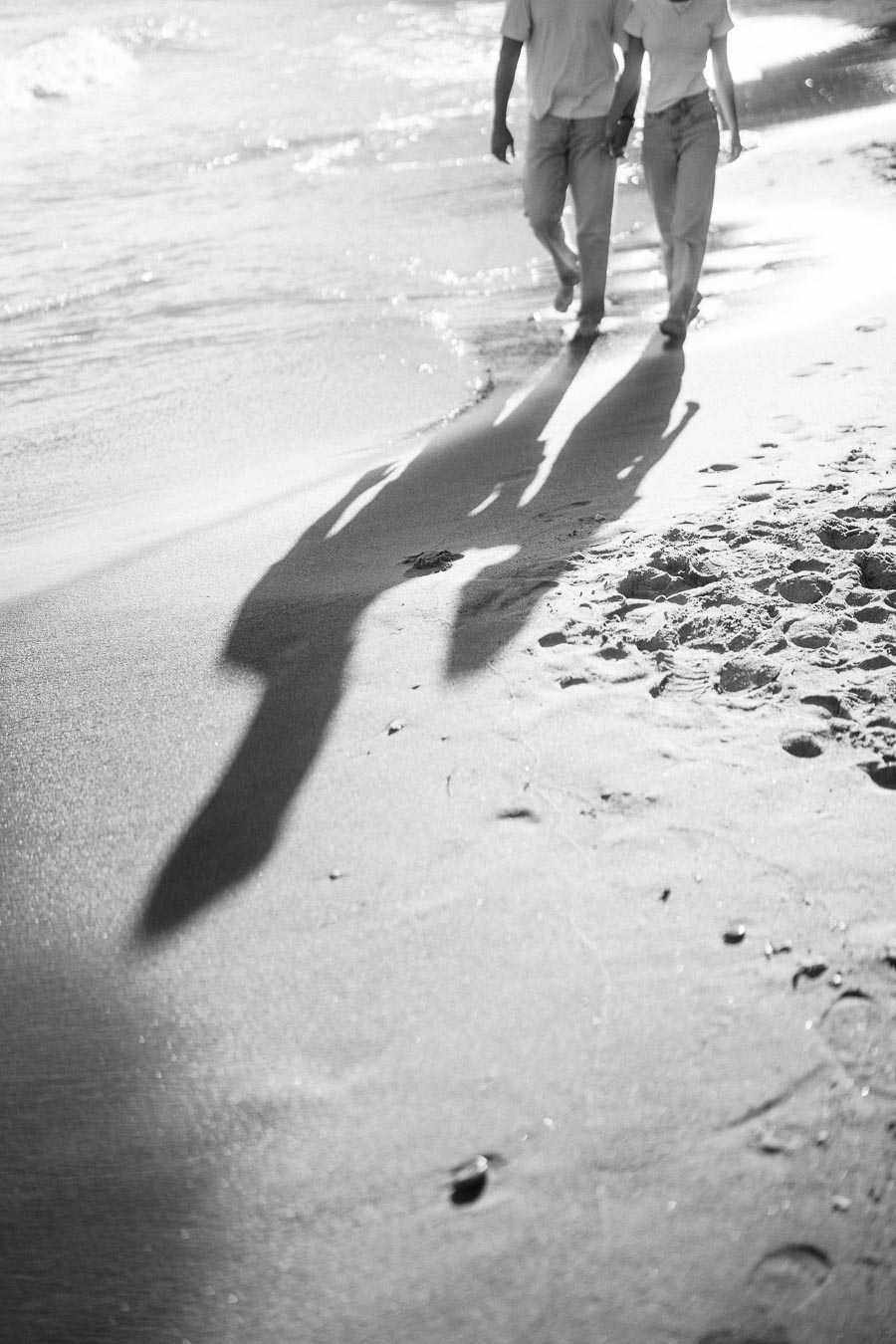 Black and white image of a couple walking hand in hand on a sandy beach, their long shadows cast on the sand by the sunlight.