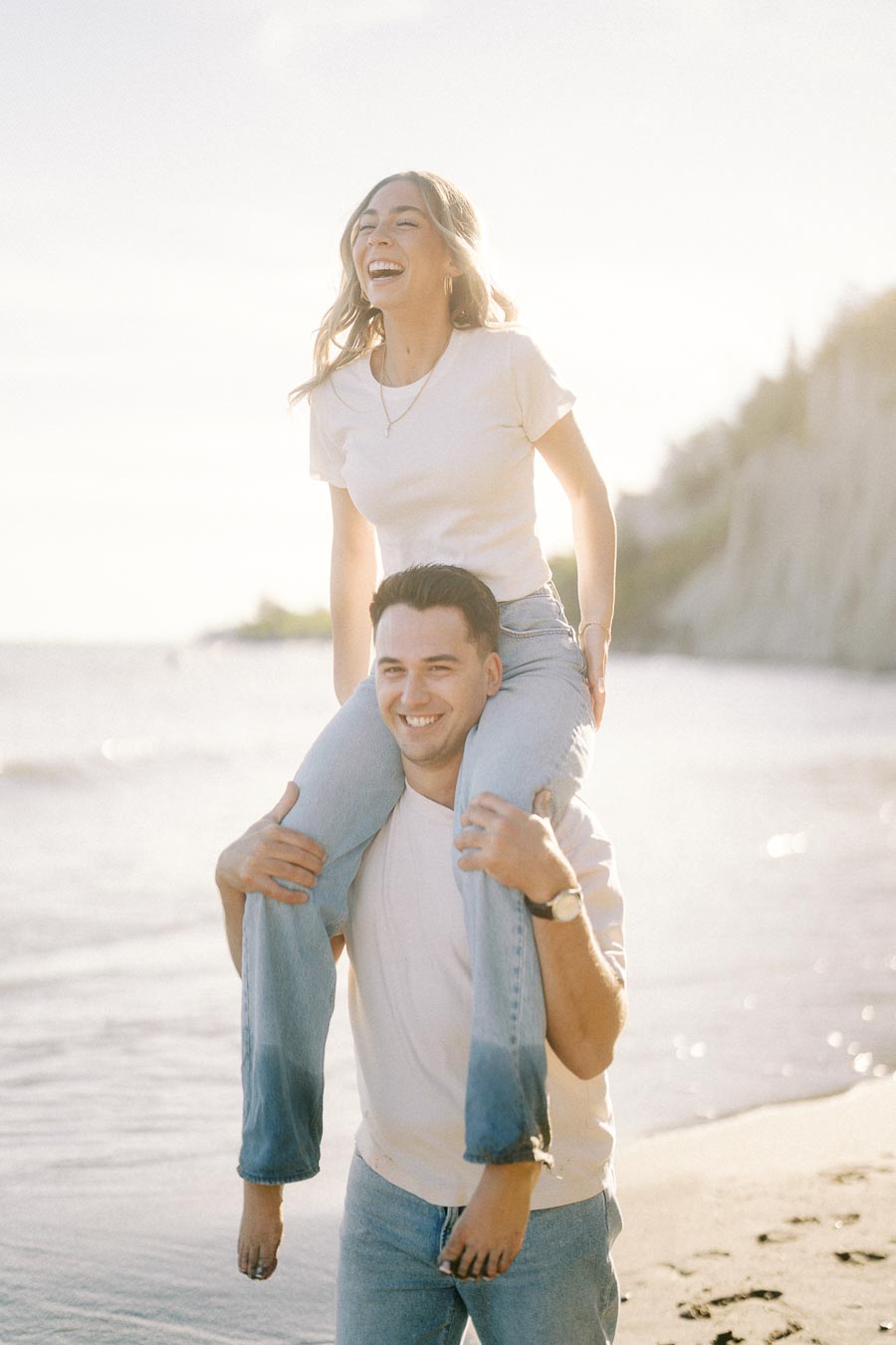 Happy couple enjoying a playful moment on the beach, with a woman sitting on a man's shoulders, both wearing casual white shirts and jeans, with ocean waves and cliffs in the background.