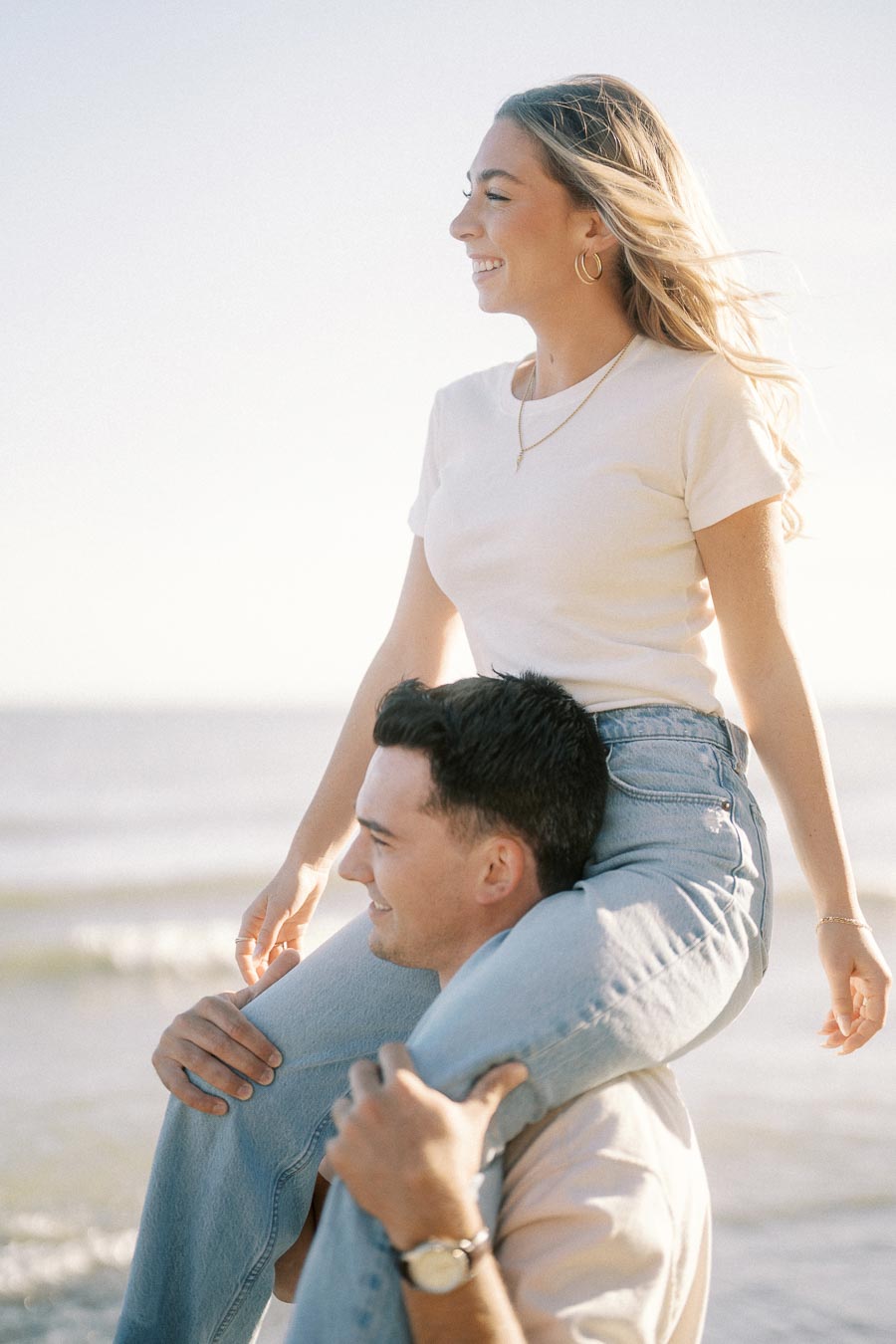 A woman smiling while sitting on a man's shoulders on a sunny beach.