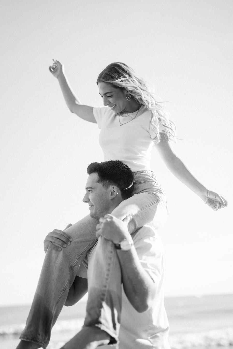 Young woman joyfully enjoying a piggyback ride on a man's shoulders at the beach, captured in black and white.