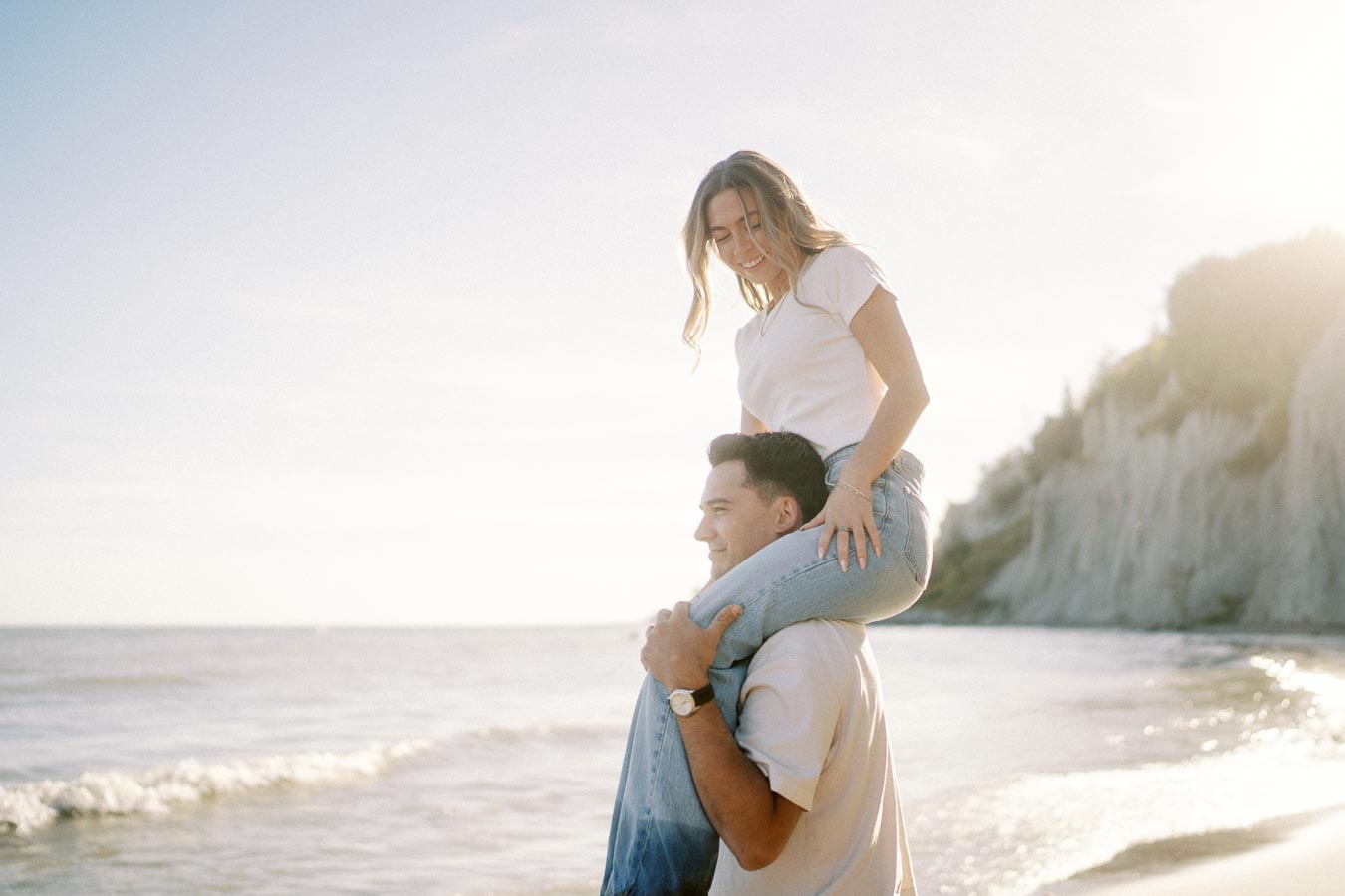 A joyful couple enjoying a sunny day at the beach, with the woman sitting on the man's shoulders, both dressed casually in jeans and white tops, with a scenic ocean and cliff background.