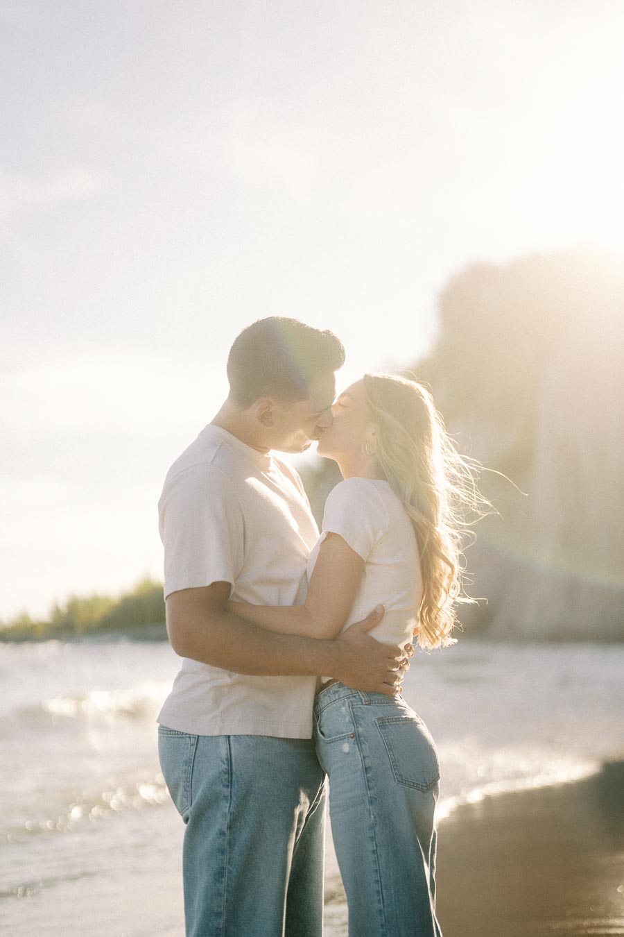 Romantic couple kissing on a sunlit beach, wearing casual white shirts and jeans, with waves gently crashing in the background.
