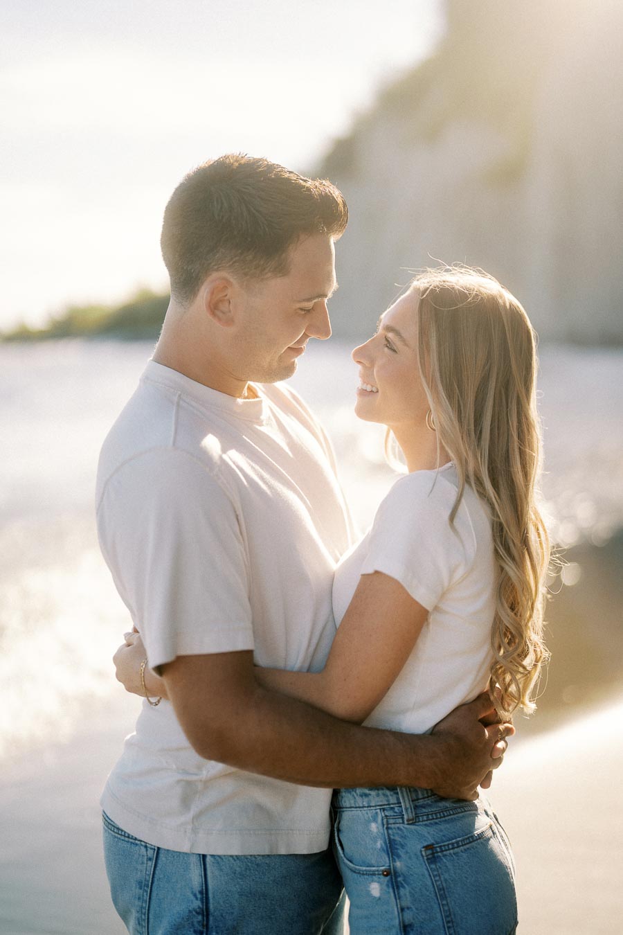 Couple embracing on a sunlit beach, wearing casual white and denim outfits, with a serene ocean backdrop and soft sunlight illuminating their faces.