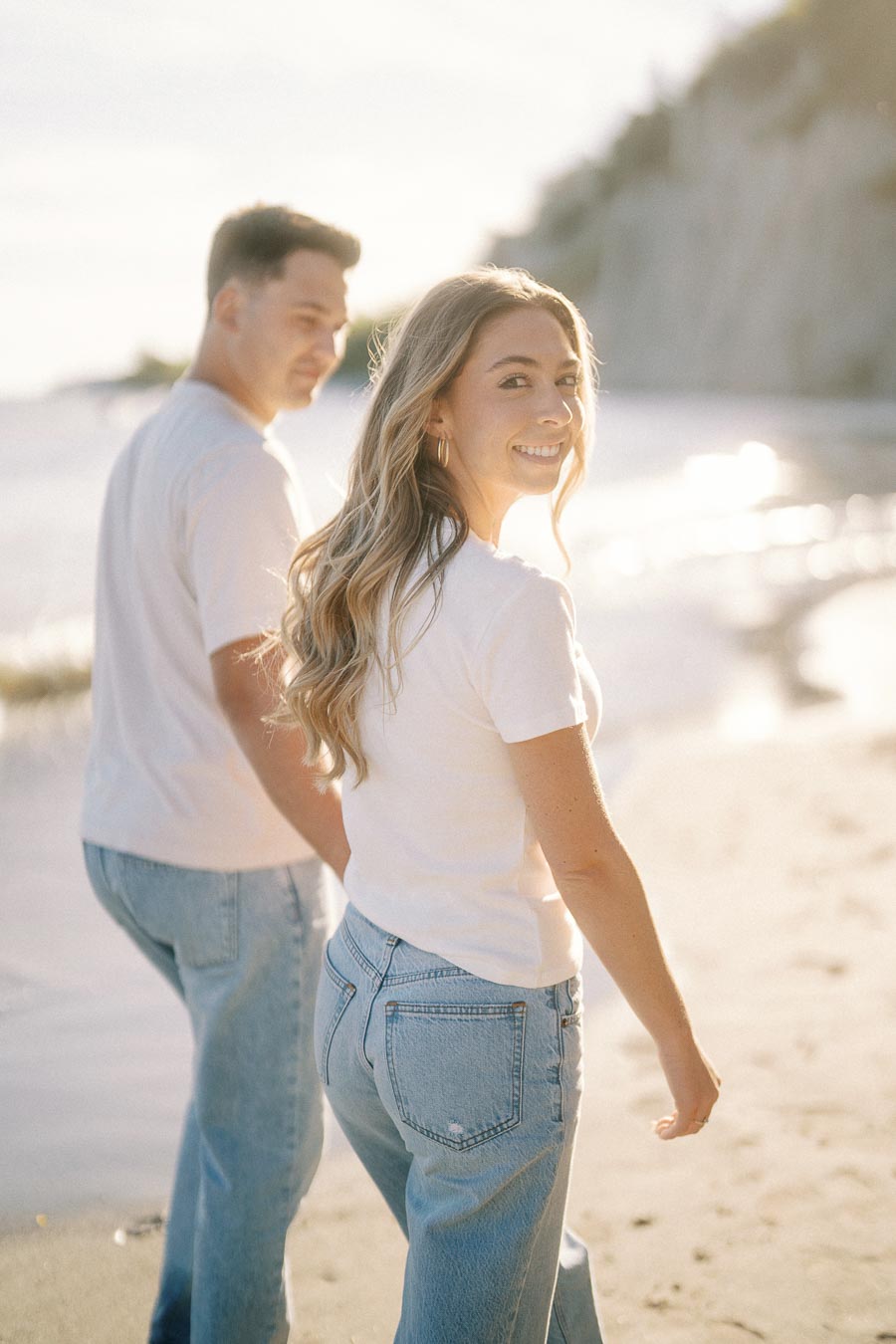 A couple walking on a sunlit beach, wearing casual white tops and jeans, with the ocean and cliffs in the background.