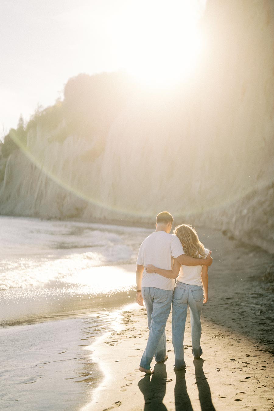 A couple walking hand in hand on a sandy beach during sunset, with cliffs in the background, wearing casual white tops and jeans, creating a romantic and serene atmosphere.