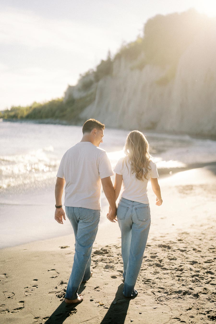 Couple holding hands walking on sandy beach at sunset, with ocean waves and cliffs in the background, wearing casual blue jeans and white tops.